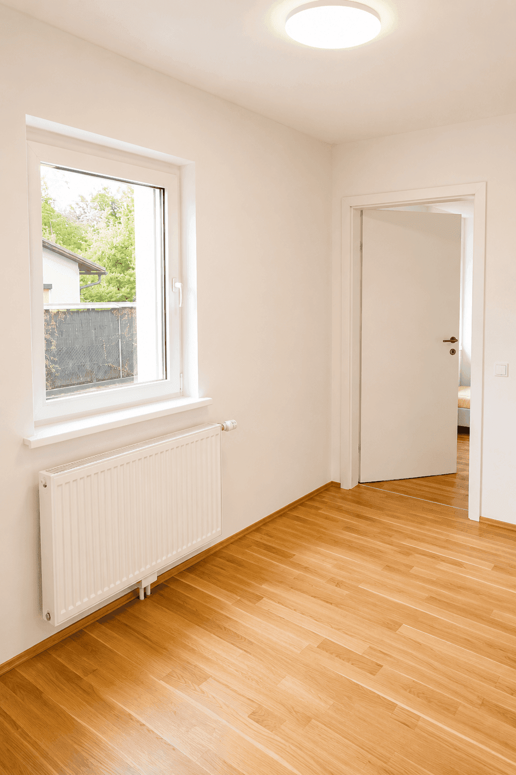 Freundliches Zimmer mit Parkettboden, Fenster und weißer Tür zu einem weiteren Raum.