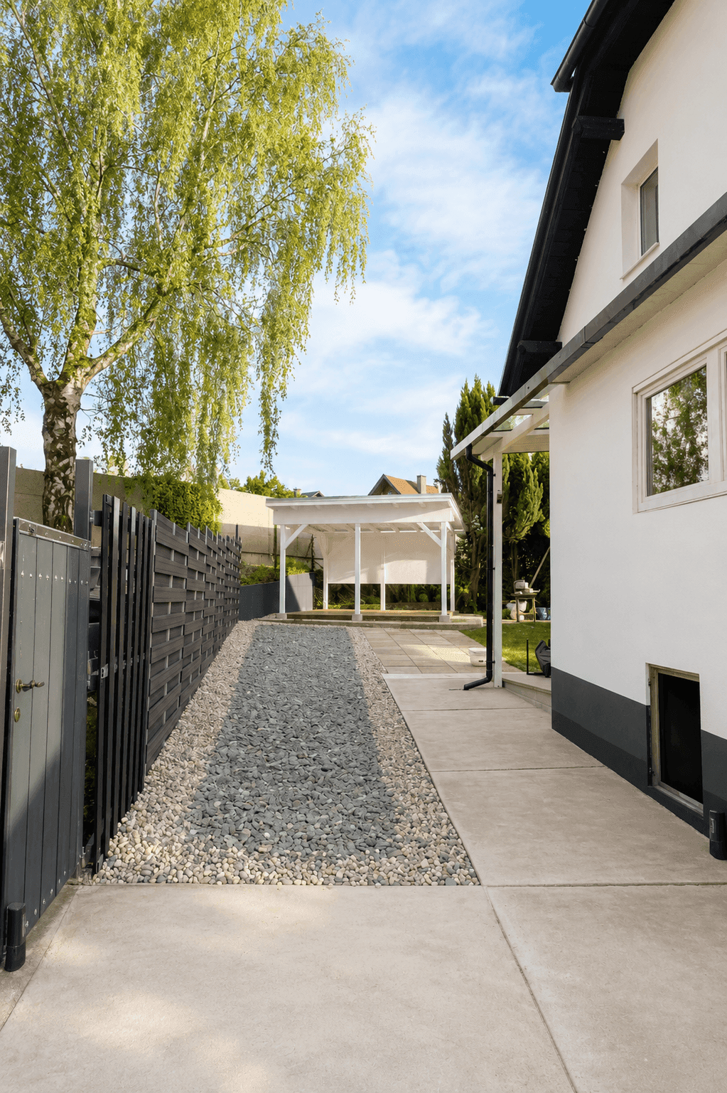 Seitlicher Zugang zum Haus mit Kiesweg, gepflegtem Garten und einer Pergola im Hintergrund.