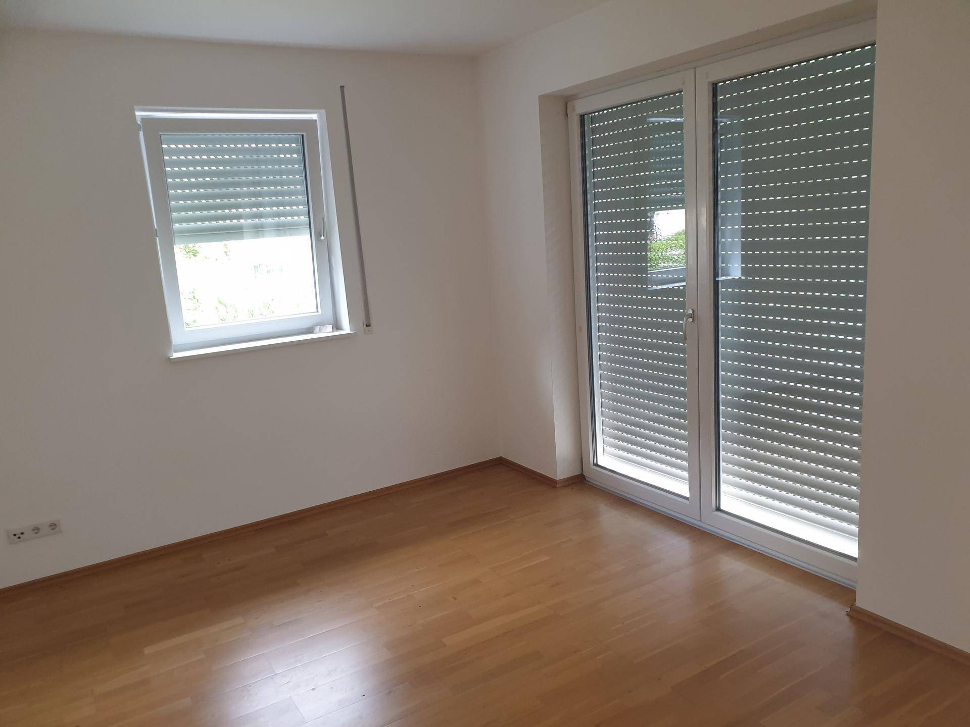 Helles Zimmer mit Parkettboden und Fenster sowie Tür mit Rollläden.