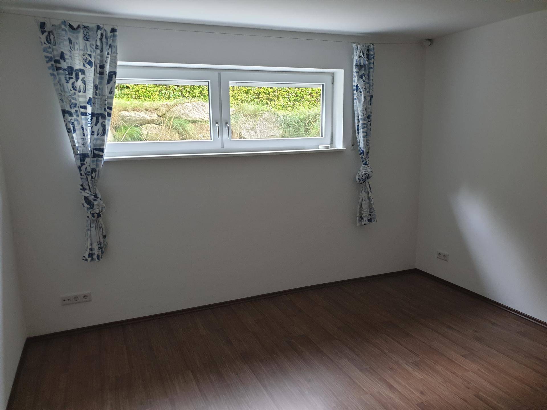 Helles Zimmer mit Laminatboden und Fenster mit Vorhängen.