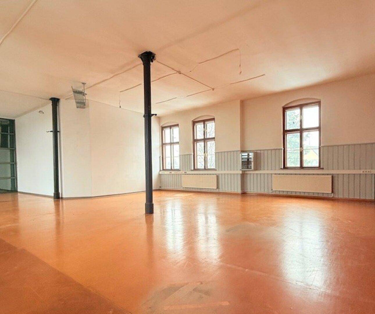 Offener Wohnbereich mit orangefarbenem Industrieboden, hohen Decken und großen Fenstern, die viel Tageslicht hereinlassen.