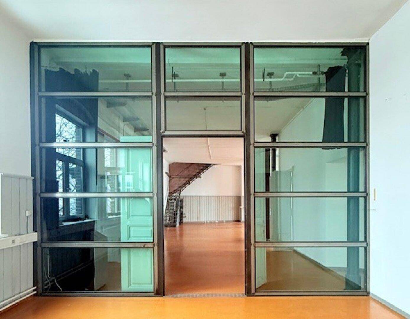 Industrielle Glaswand mit Tür, die einen Durchgang zu einem weiteren offenen Bereich ermöglicht, mit sichtbaren Leitungen.