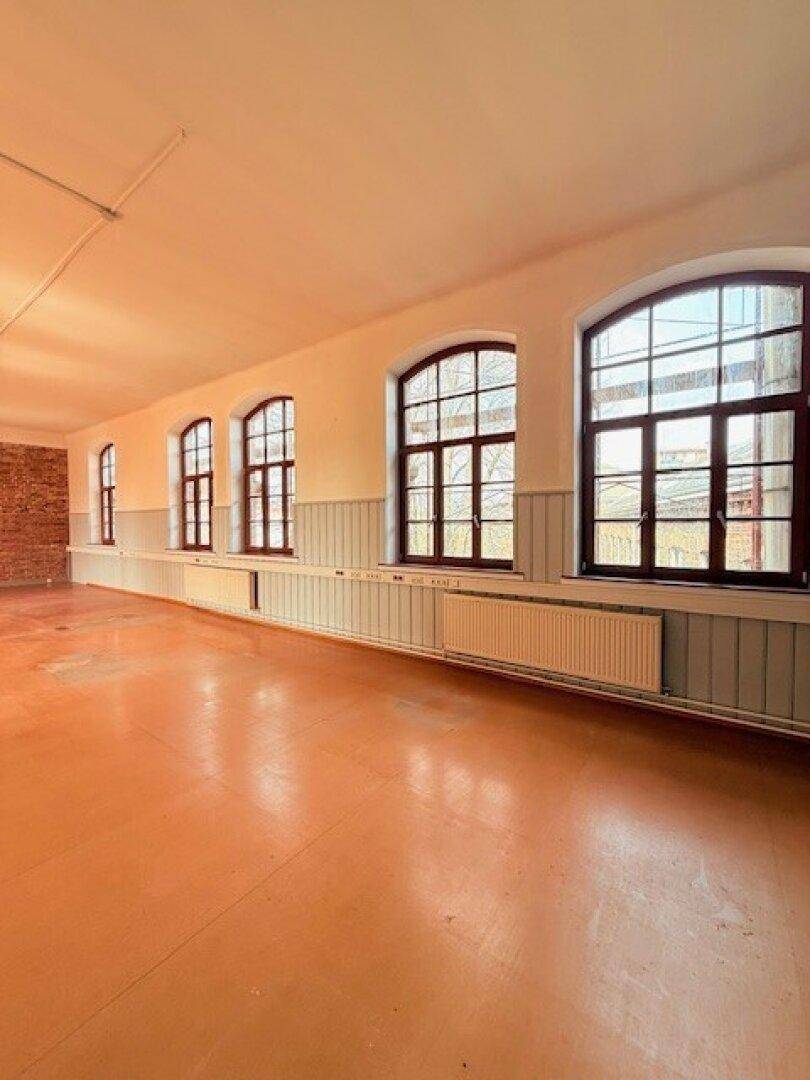 Heller, weitläufiger Raum mit mehreren Bogenfenstern und Heizkörpern unter einer Ziegelwand, bietet viel natürliches Licht.