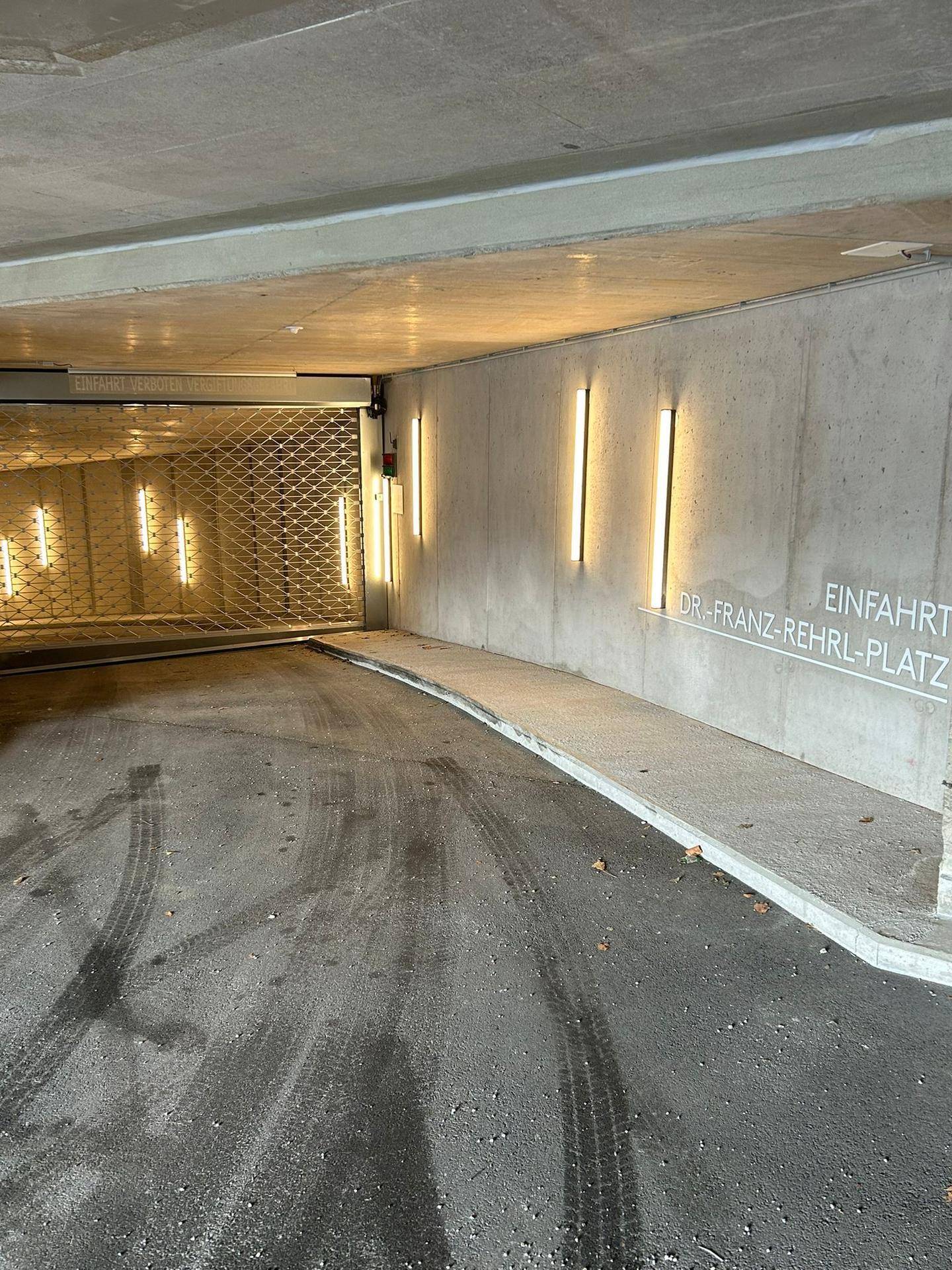 Einfahrt zu einer Tiefgarage mit Rampe, Rolltor und beleuchteter Betonwand.
