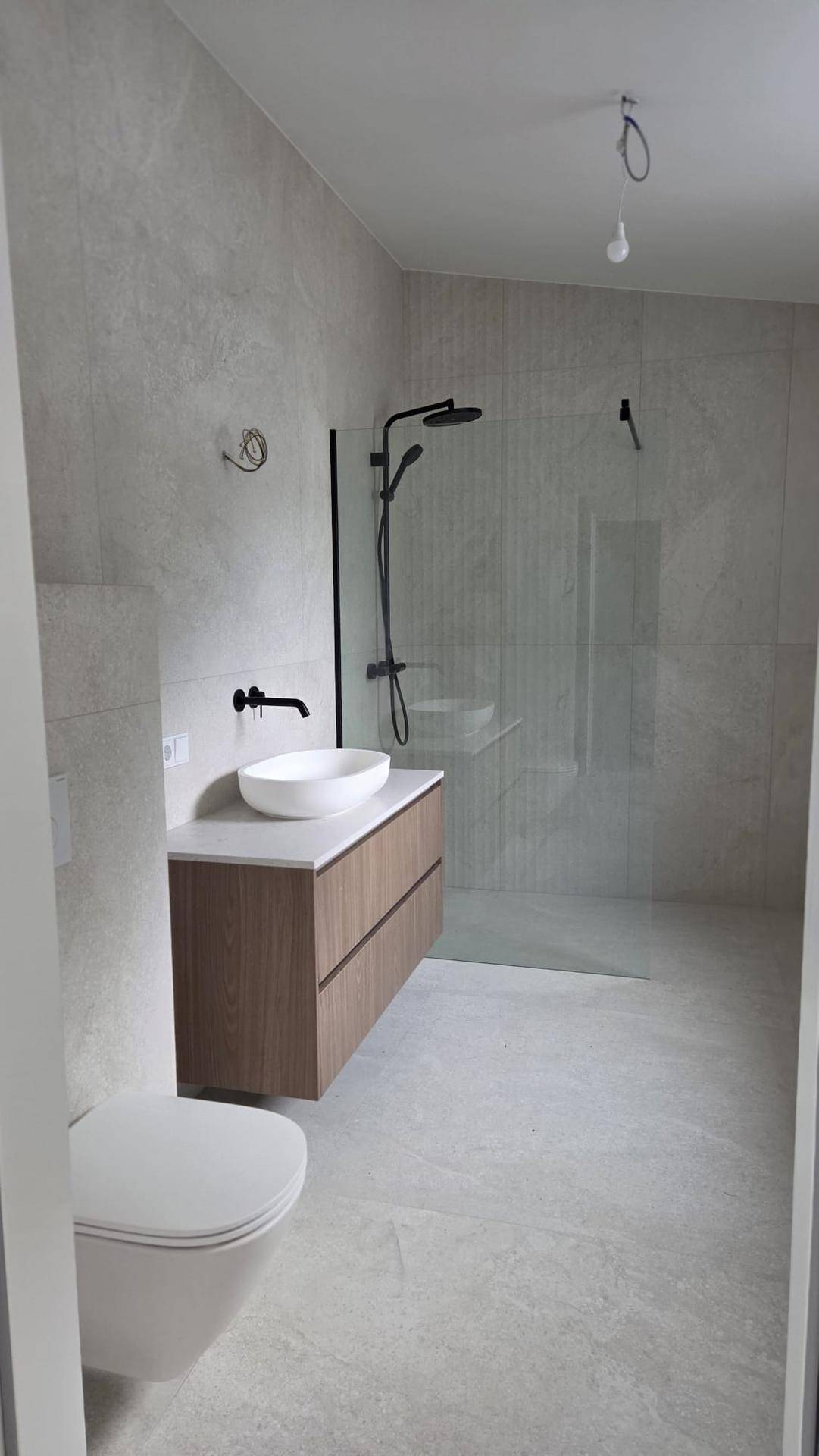 Modernes Badezimmer mit hellgrauen Fliesen, wandhängendem WC, Einzelwaschbecken und begehbarer Dusche.