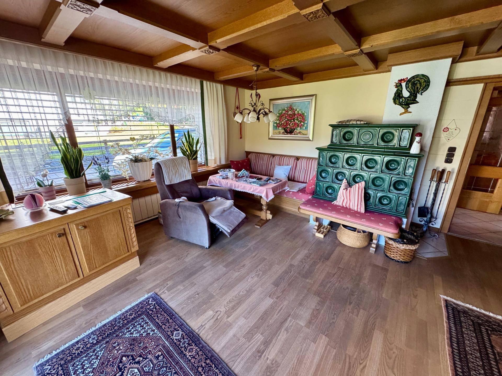Gemütliches Wohnzimmer mit traditionellem Kachelofen, Holzbalkendecke und großen Fenstern.
