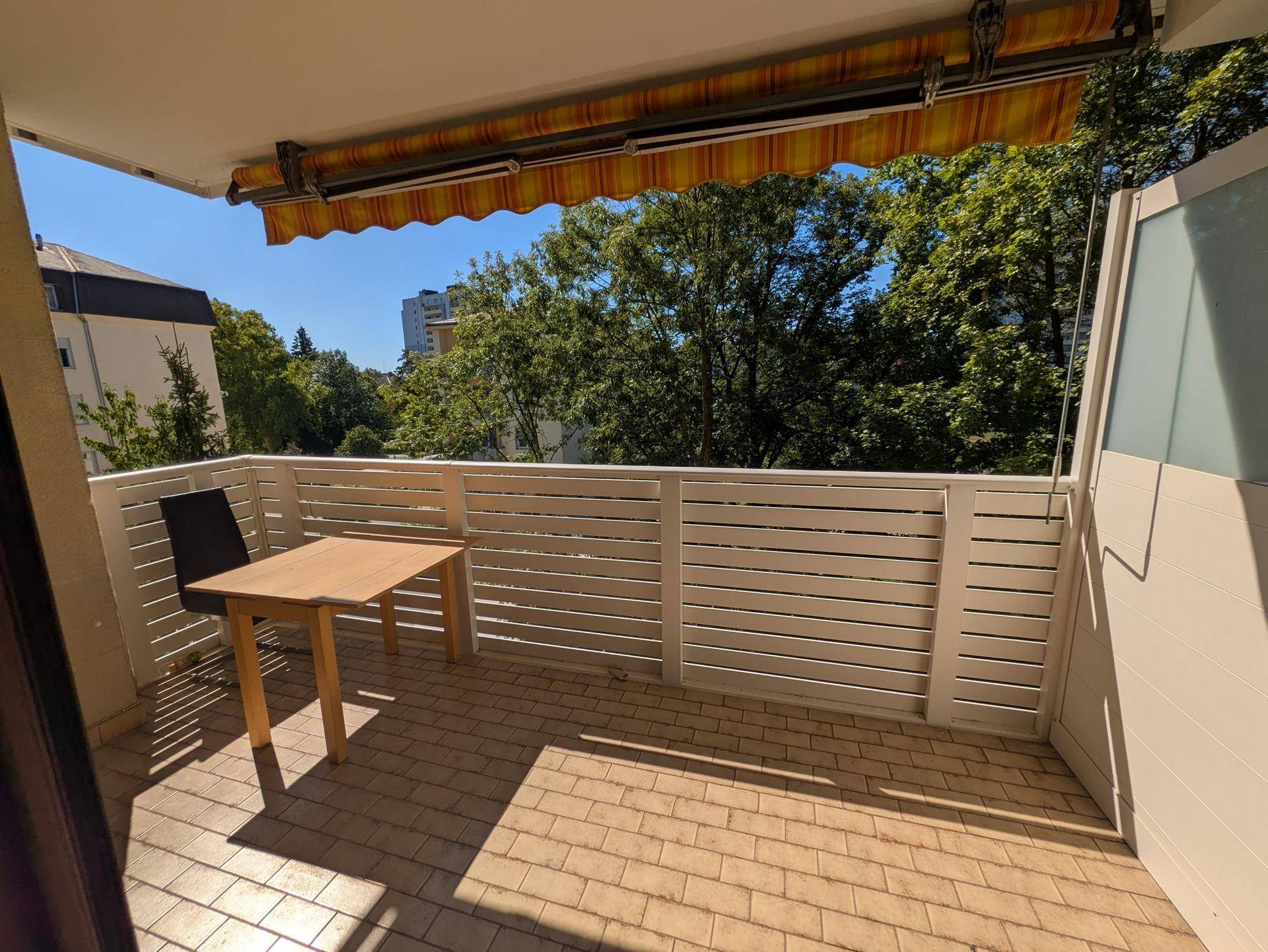 Sonniger Balkon mit Fliesenboden, einer Markise und Blick ins Grüne mit Stadtgebäuden im Hintergrund.