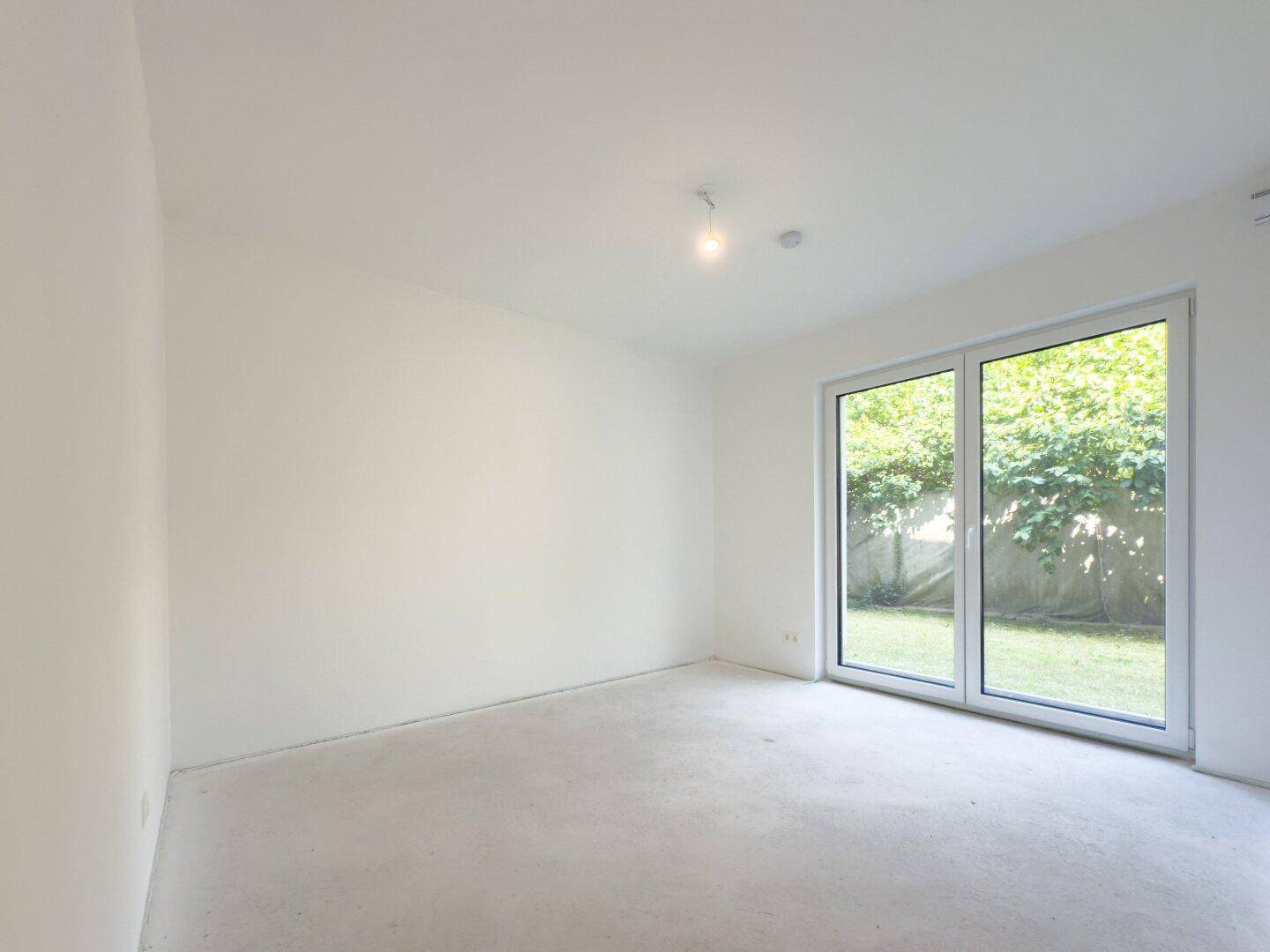 Unmöblierter Raum mit großer Fenstertür und direktem Blick auf die grüne Außenanlage.