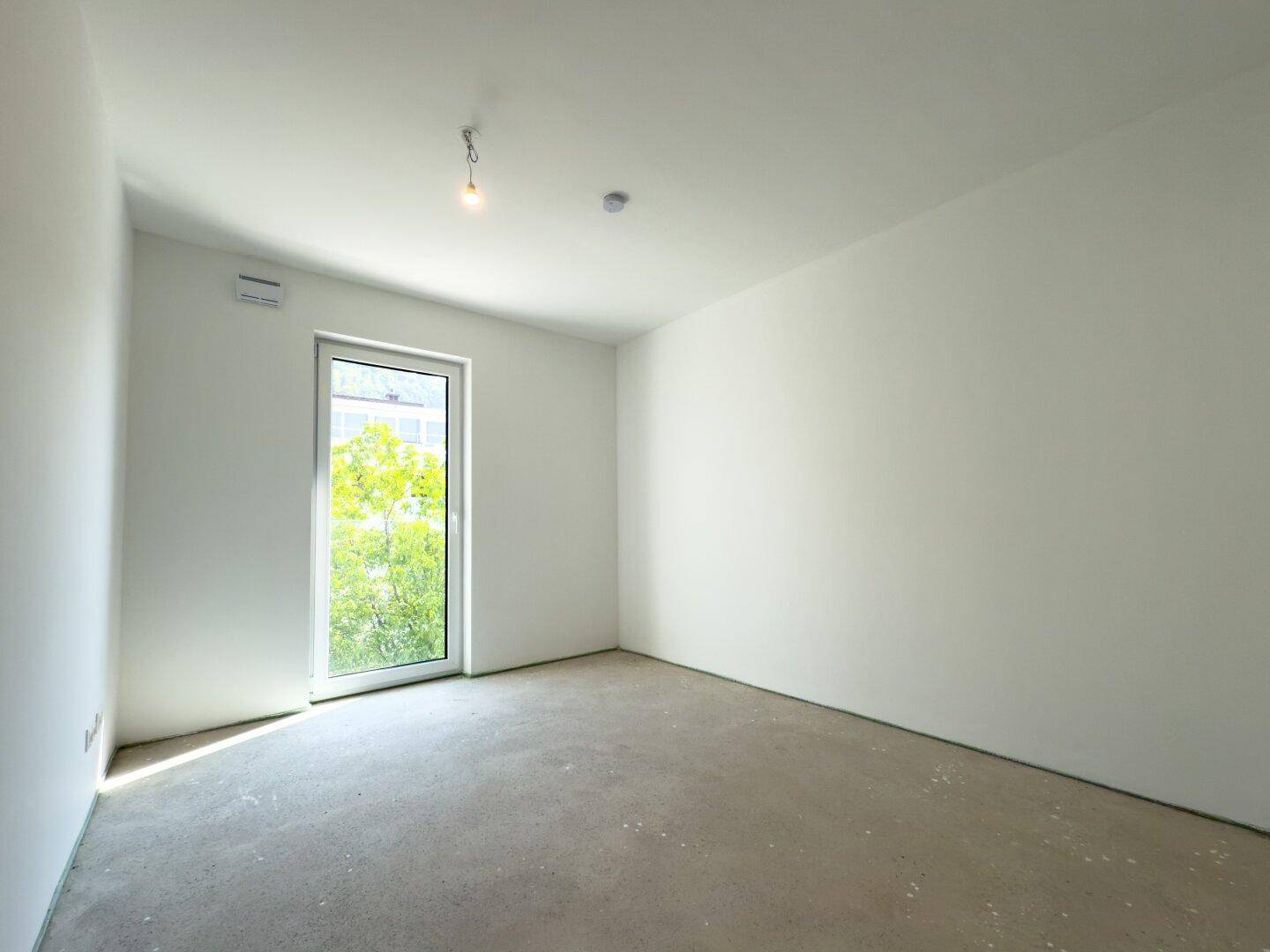 Helles, unmöbliertes Schlafzimmer im Rohbauzustand mit Fenster und Zugang ins Freie.