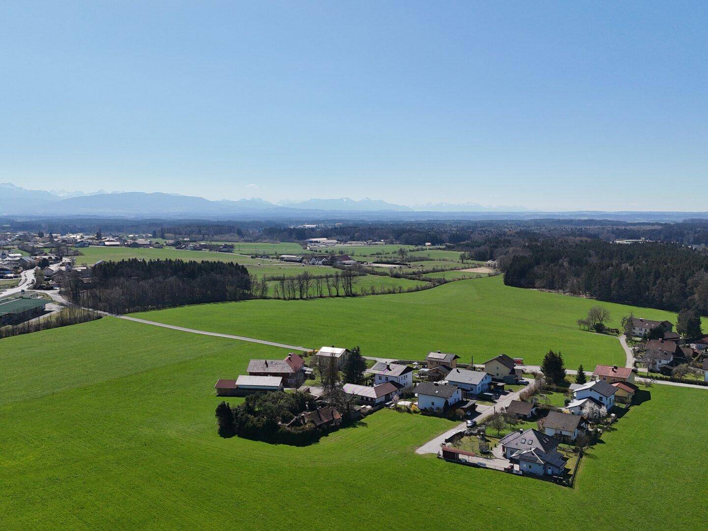 Panoramische Luftaufnahme einer ländlichen Siedlung mit weiten Feldern und Bergen am fernen Horizont.