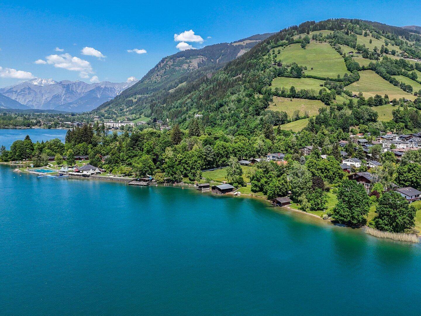 Luftaufnahme eines malerischen Sees mit Häusern am Ufer, umgeben von grünen Hügeln und Bergen.