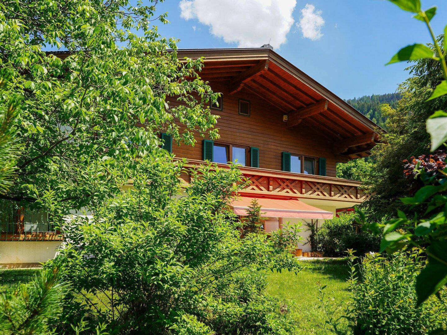 Traditionelles Holzhaus mit Holzbalkonen, umgeben von üppigem Grün und einem roten Sonnenschutz.