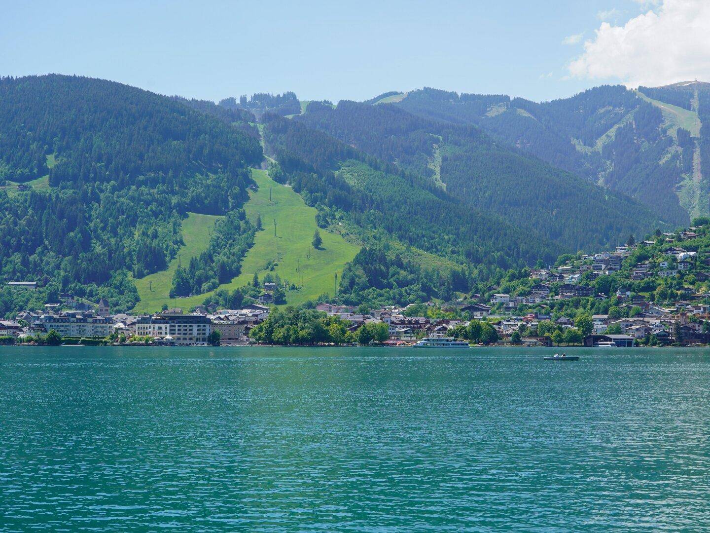 Stadtansicht am Seeufer mit grünen Bergen und Skipisten im Hintergrund unter klarem Himmel.