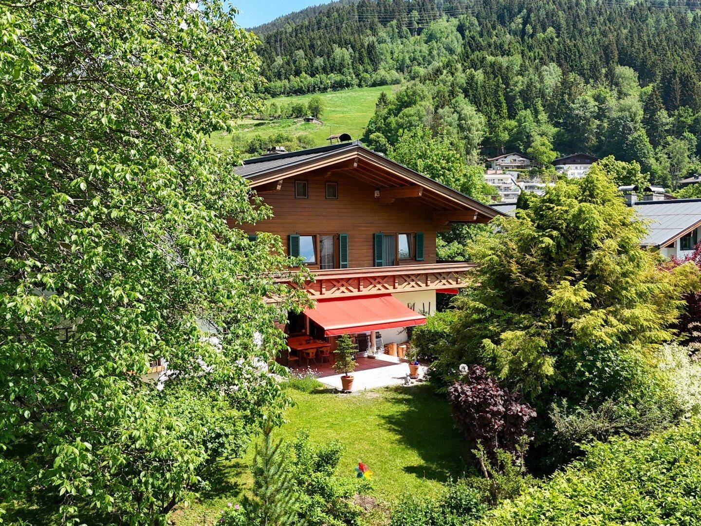 Traditionelles Holzhaus mit Garten, roter Markise und grünen Hügeln im Hintergrund unter blauem Himmel.