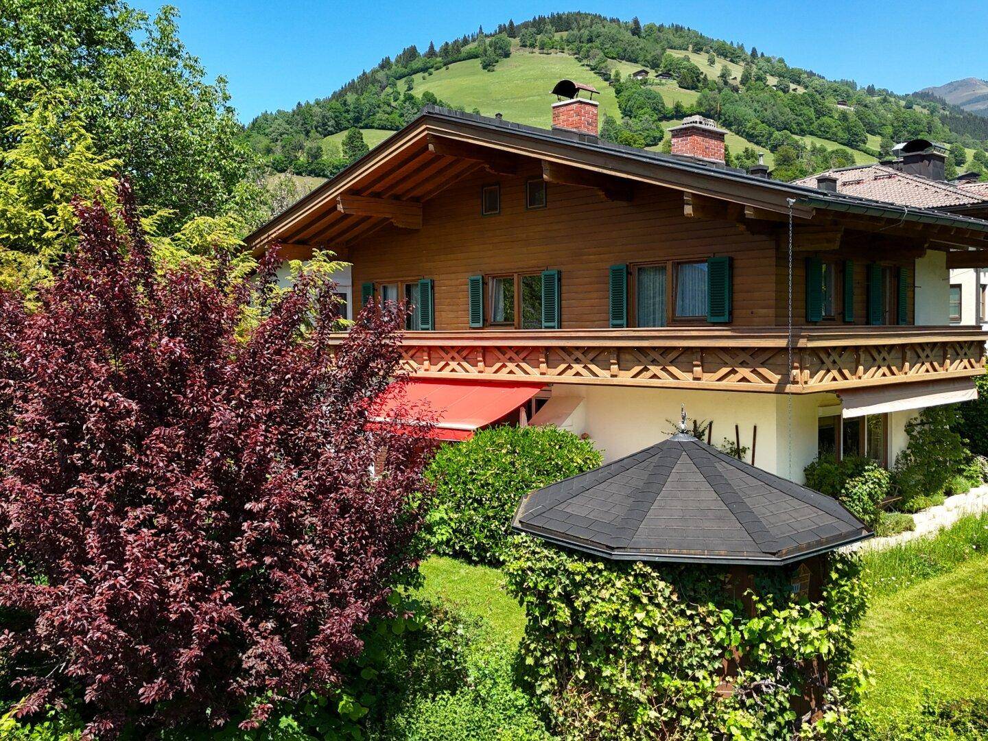 Traditionelles Holzhaus mit roter Markise und einem von Efeu bewachsenen Pavillon vor grünen Hügeln.