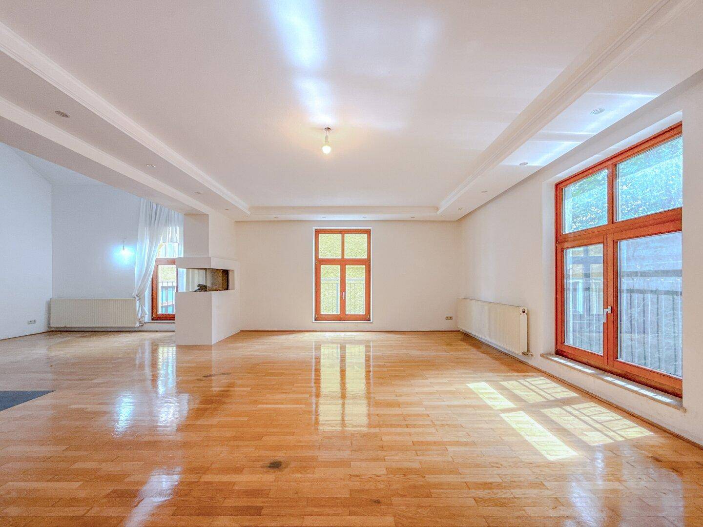 Heller, leerer Wohnraum mit Holzboden, Kamin und großen Fenstern für viel Tageslicht.