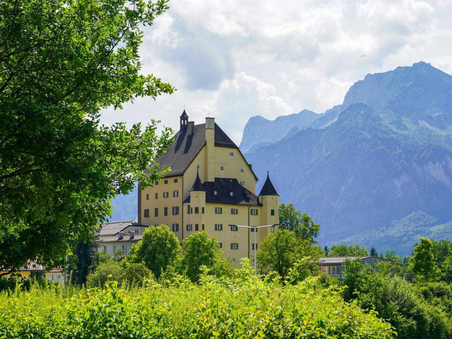 Beeindruckendes historisches Gebäude mit Türmen, umgeben von grüner Natur und majestätischen Bergen.