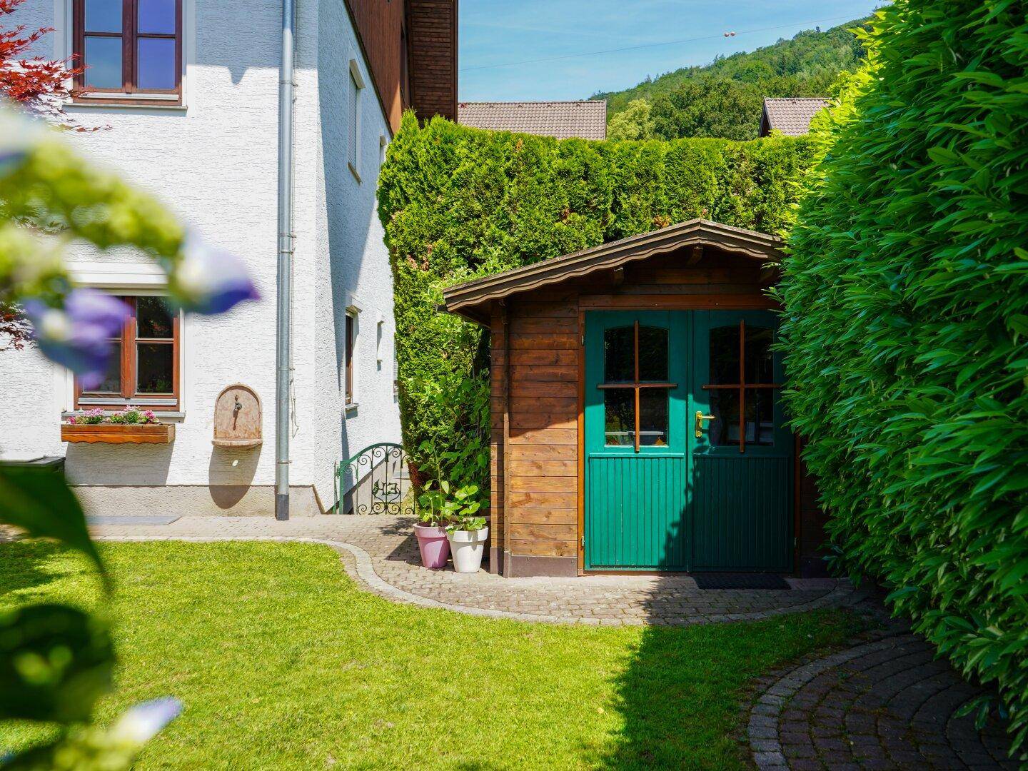Idyllischer Gartenbereich mit einem charmanten Holzschuppen, umgeben von einer hohen Hecke und Bergblick.