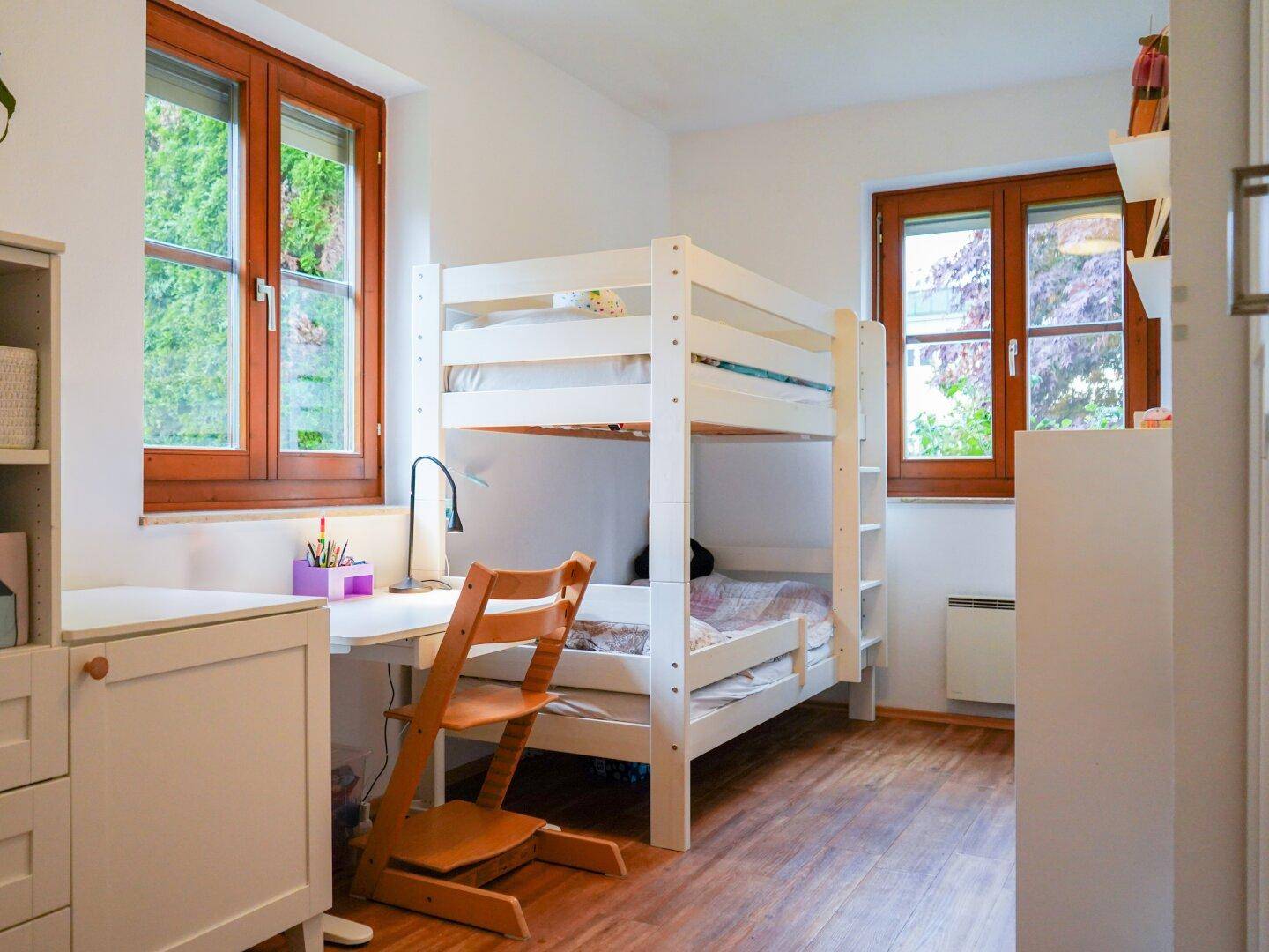Kinderzimmer mit Etagenbett, Schreibtisch und Holzboden, hell durch zwei große Fenster.