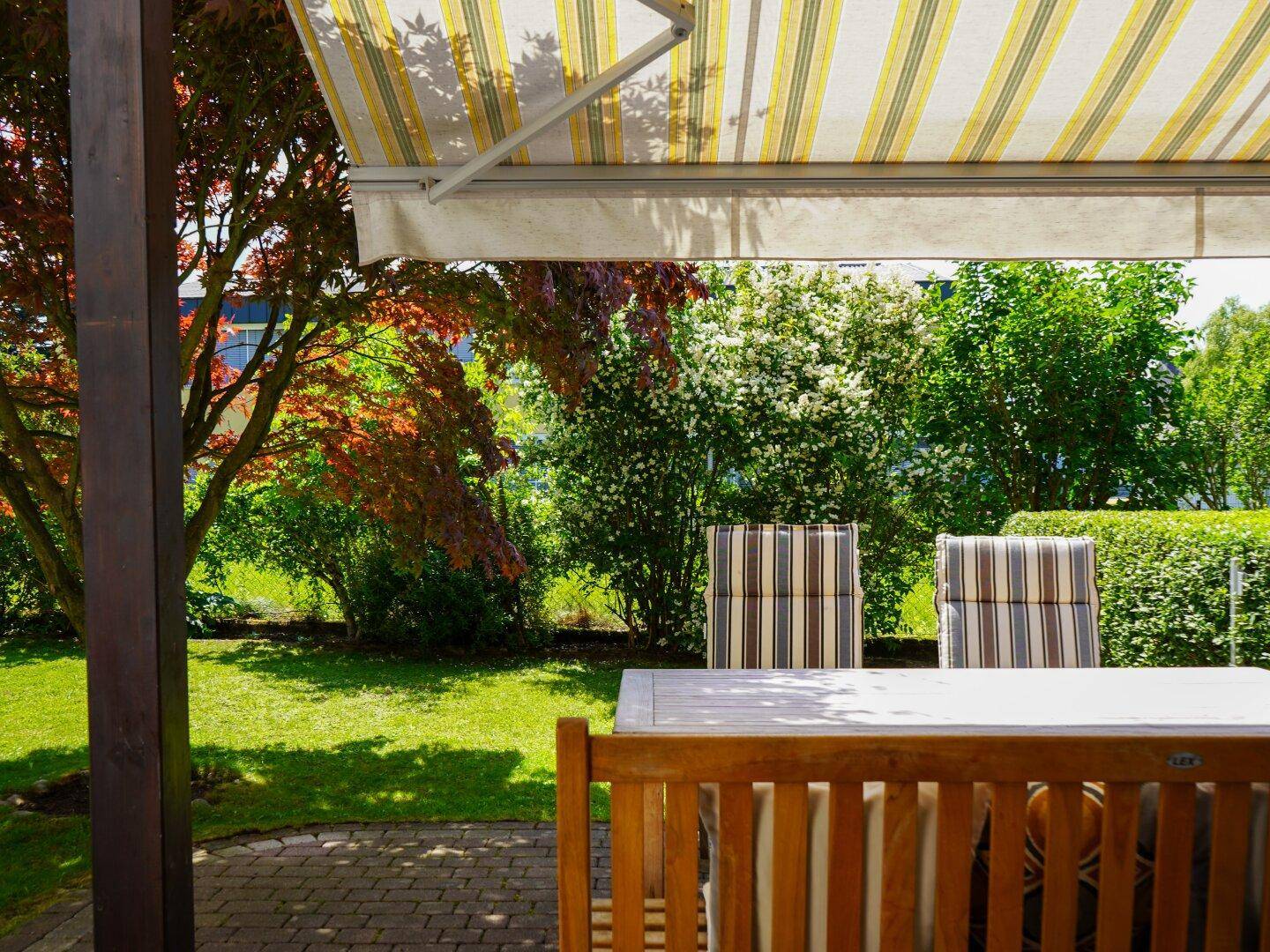 Blick von der überdachten Terrasse in den gepflegten Garten mit üppigem Grün und blühenden Sträuchern.