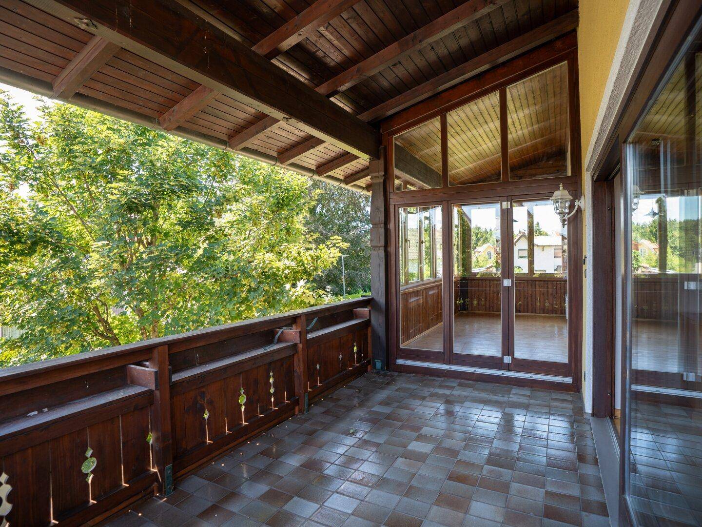 Überdachter Balkon mit Fliesenboden und Holzbalken, angrenzend an einen Wintergarten.
