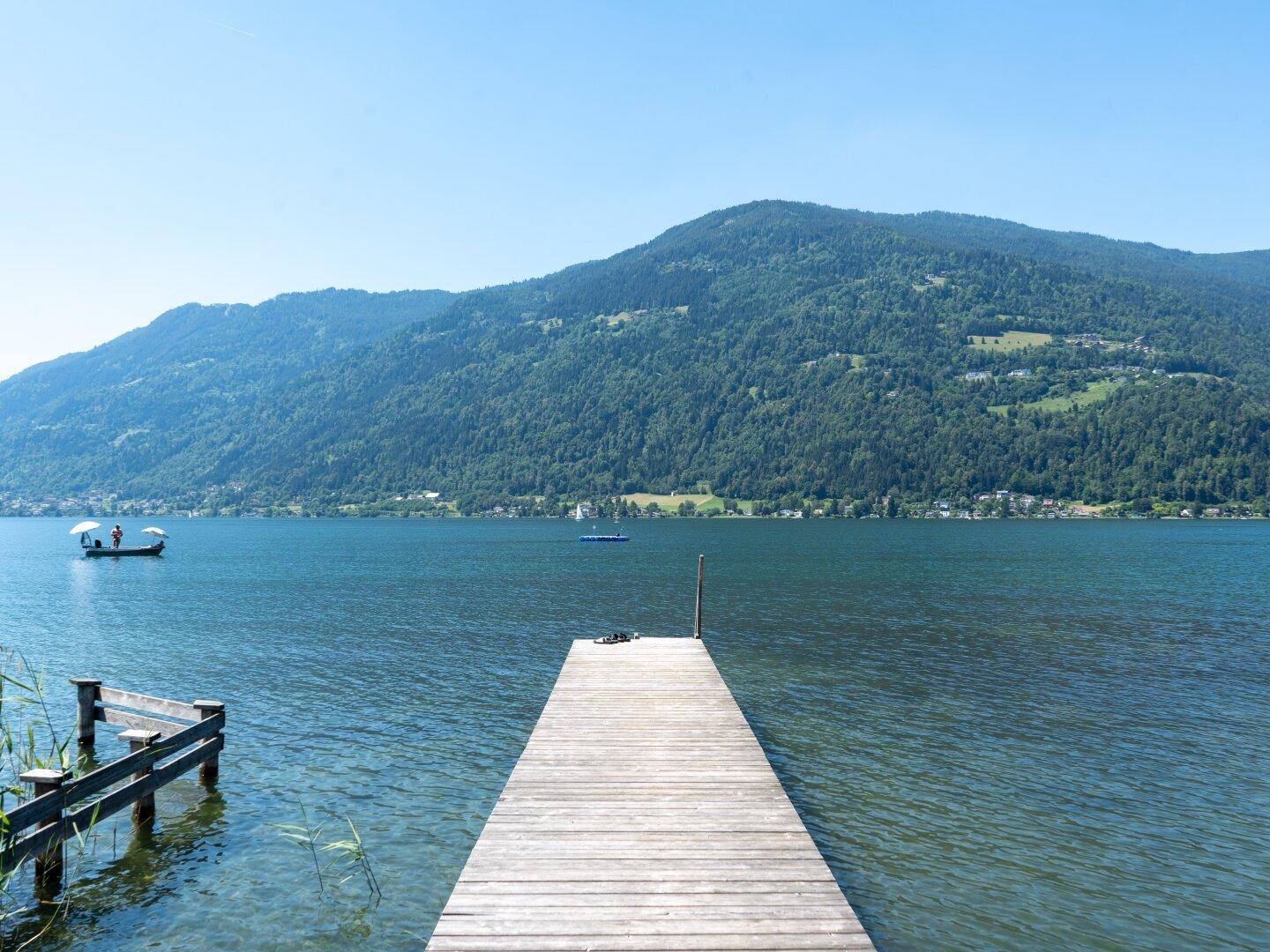 Langer Holzsteg führt in den klaren See, umgeben von grünen Bergen unter blauem Himmel.
