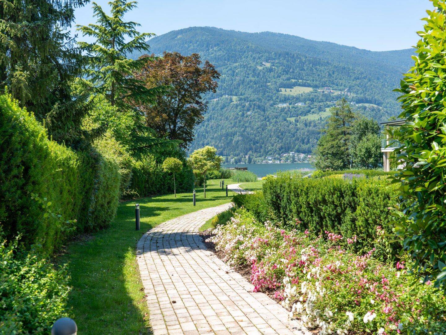 Geschwungener Gartenweg durch blühende Beete, führt zu einem malerischen Seeblick mit Bergen.