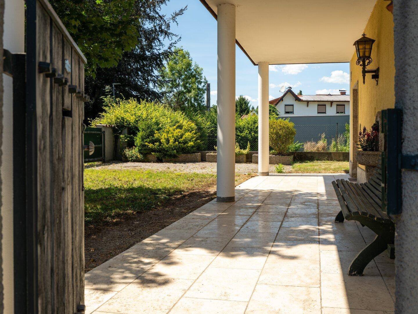 Überdachte Terrasse mit Fliesenboden und Holzsitzbank, angrenzend an einen grünen Gartenbereich.