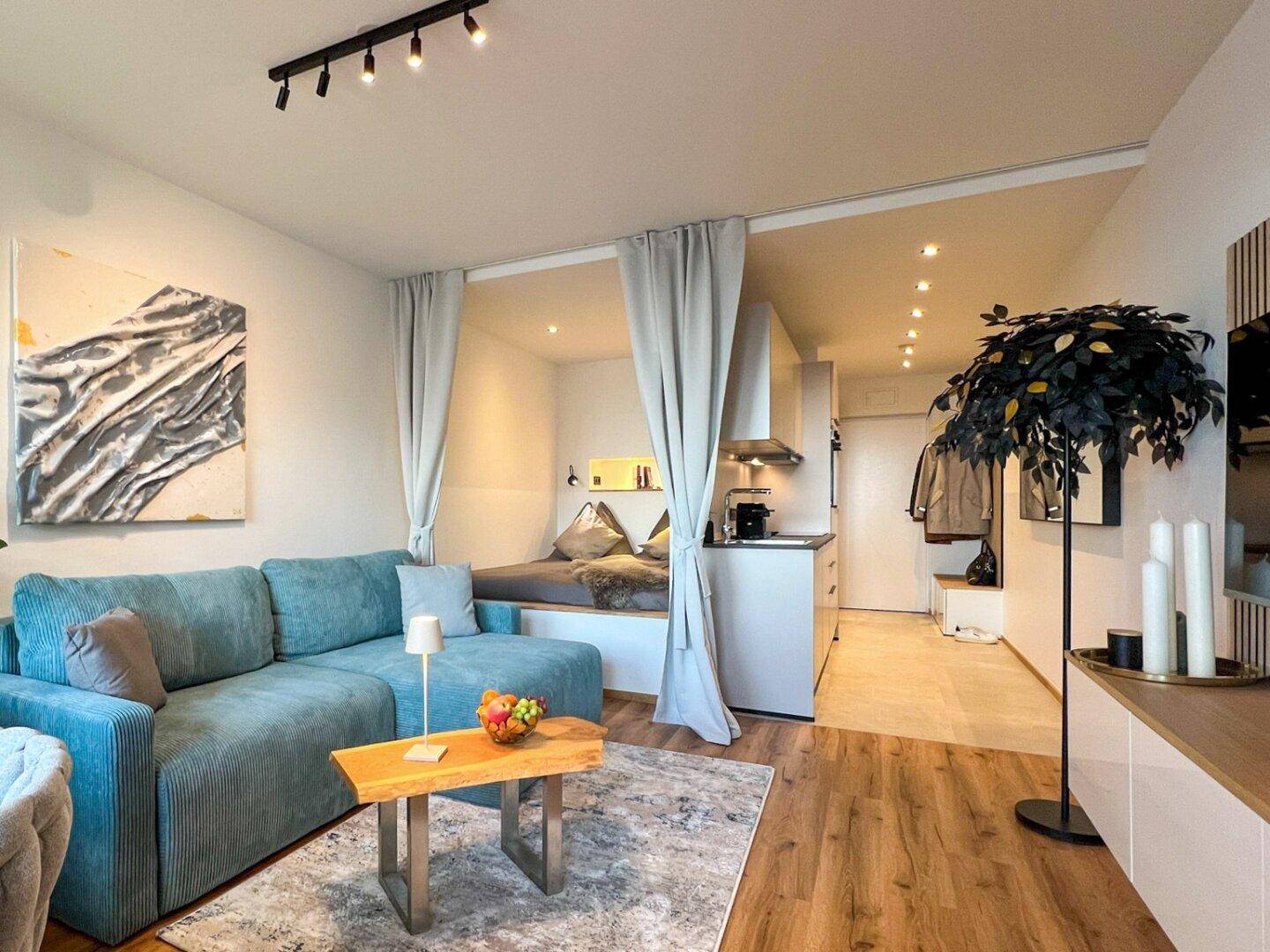 Offener Wohnbereich mit blauer Couch, Holzboden und integrierter Küchenzeile.