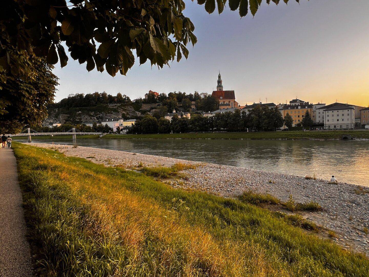 Malerische Flusslandschaft mit Uferpromenade, Bäumen und historischer Stadtkulisse im Abendlicht.