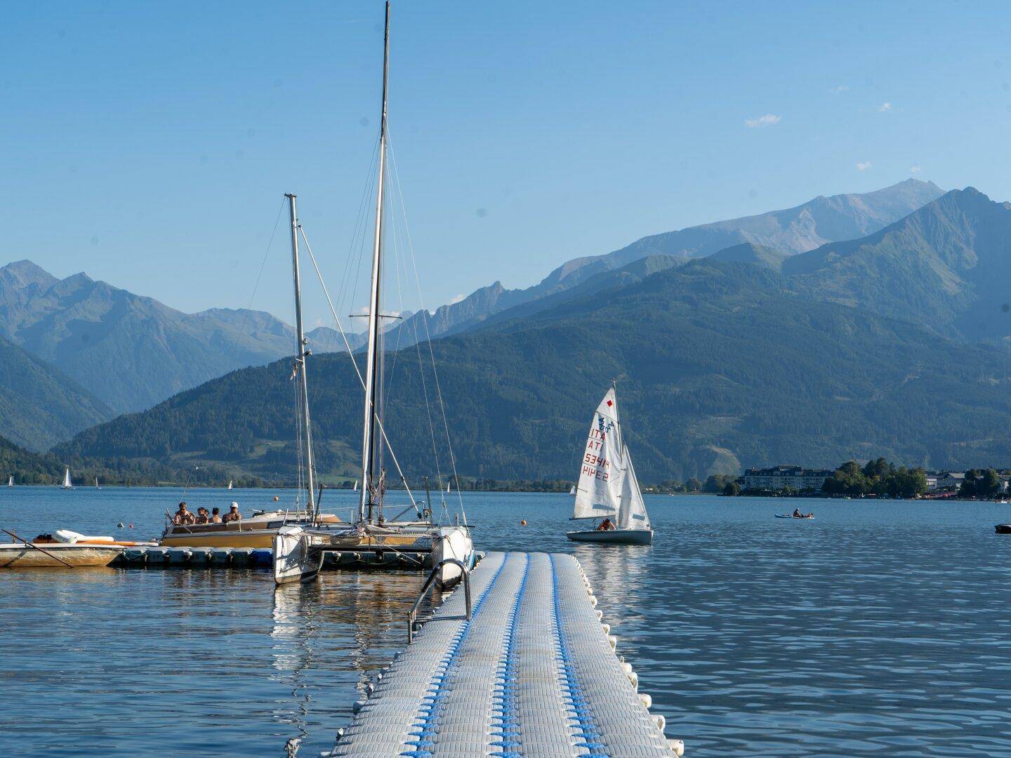 Panoramablick auf einen See mit Segelbooten, einem Steg und majestätischen Bergen im Hintergrund unter blauem Himmel.