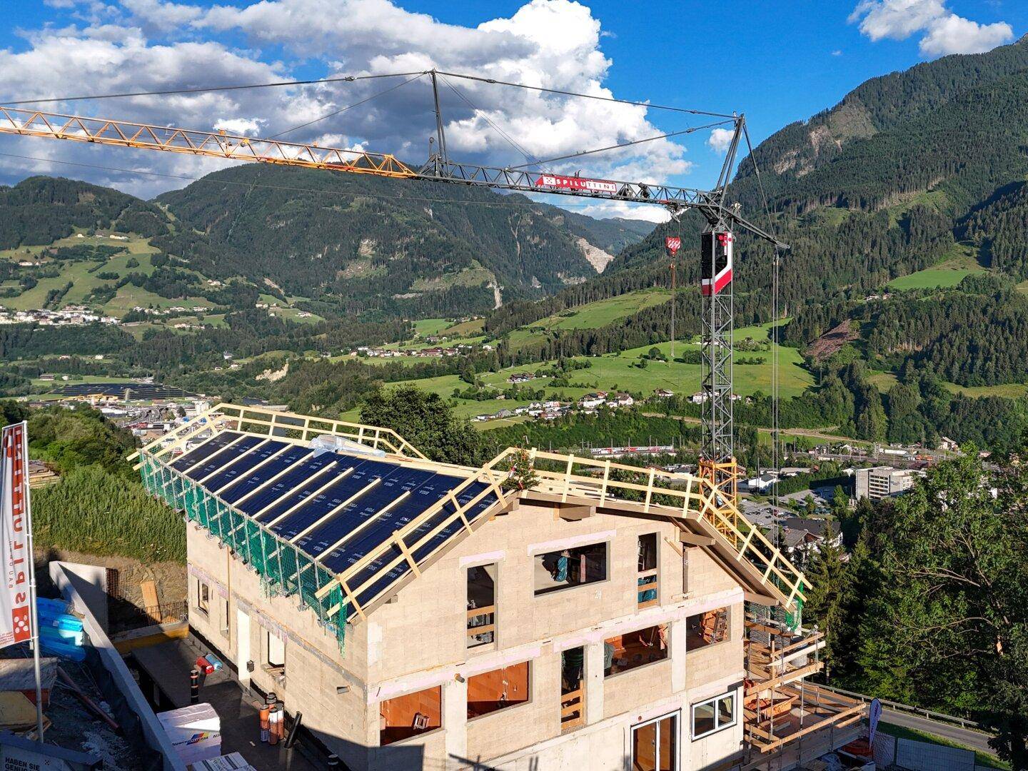 Neubau eines Hauses im Bau mit Kran und Solarpaneelen auf dem Dach, umgeben von einer Berglandschaft.