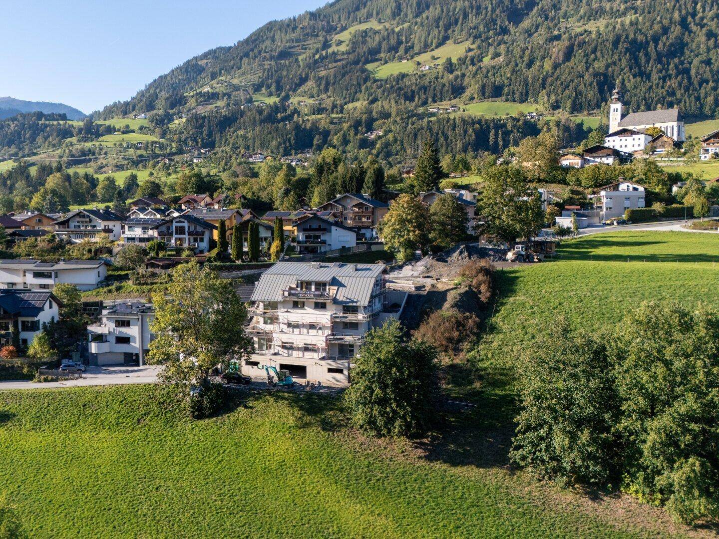 Neubauprojekt in alpiner Umgebung, umgeben von grünen Hügeln und einem Dorf.