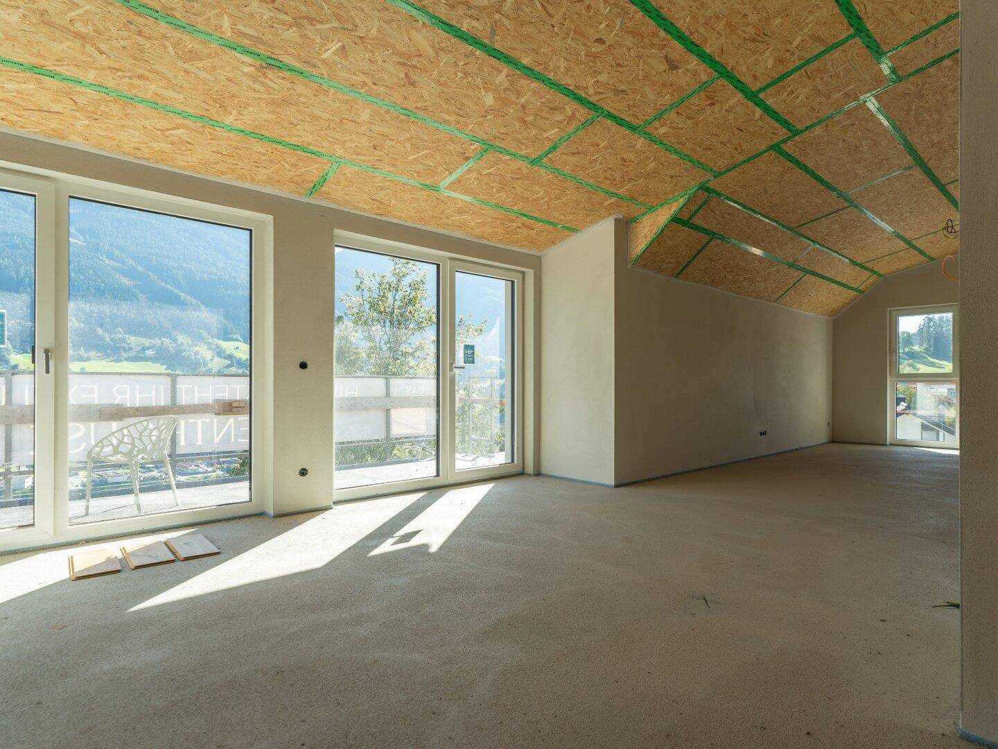 Unfertiger Raum mit schrägen Decken, großen Fenstern und Blick auf die Berge.