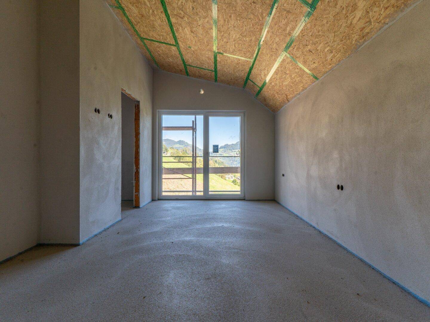 Unfertiger Raum mit schräger Decke und einer großen Fenstertür, die ins Freie führt.