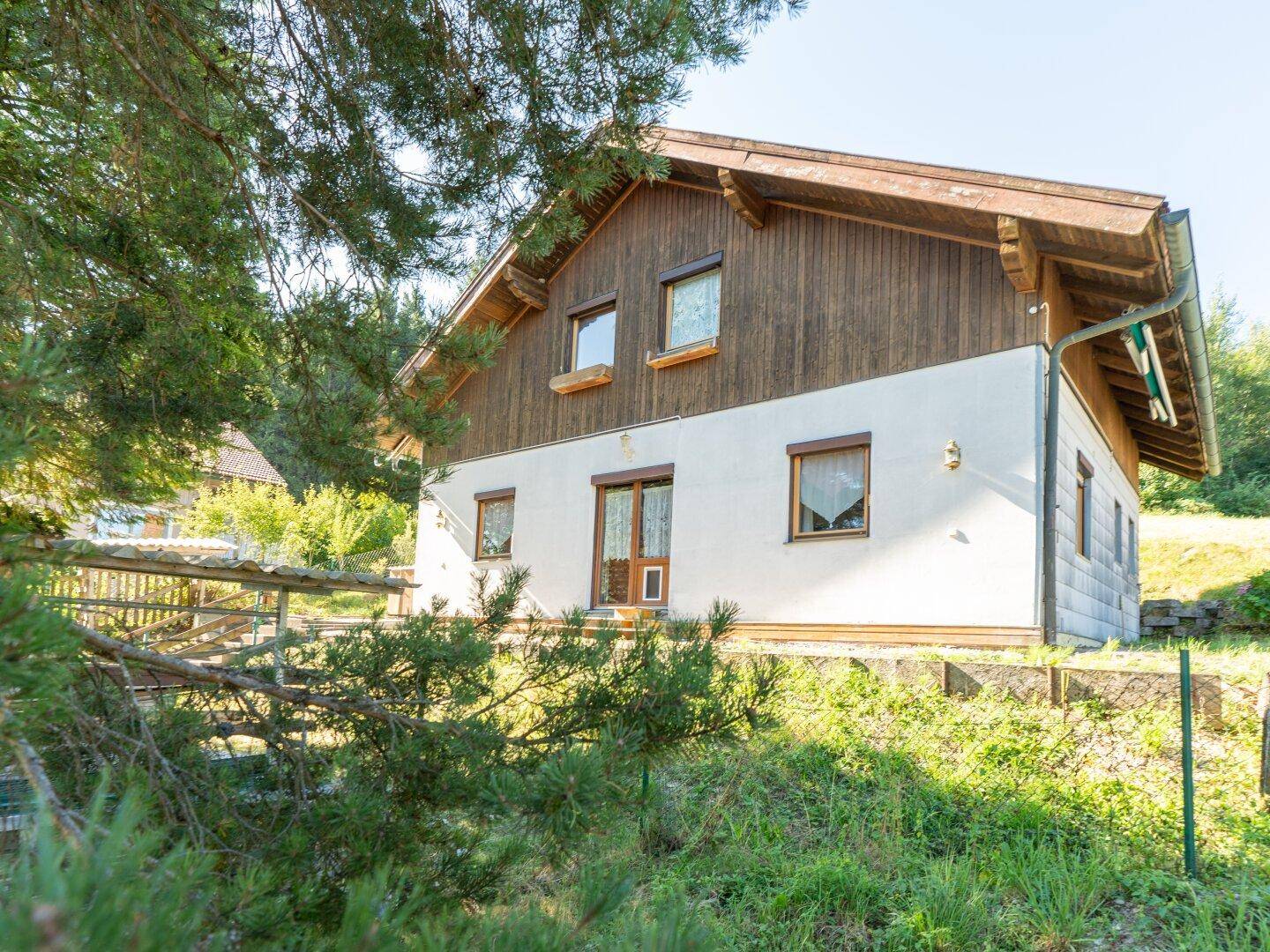 Älteres Einfamilienhaus mit Holzfassade und weißem Putz, umgeben von viel Grün und Bäumen.