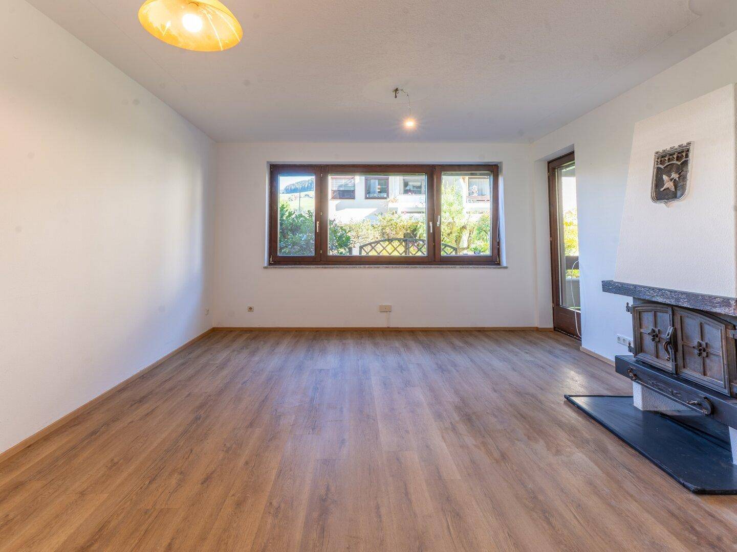 Leeres Wohnzimmer mit Holzboden, Kamin und großen Fenstern für viel Tageslicht.