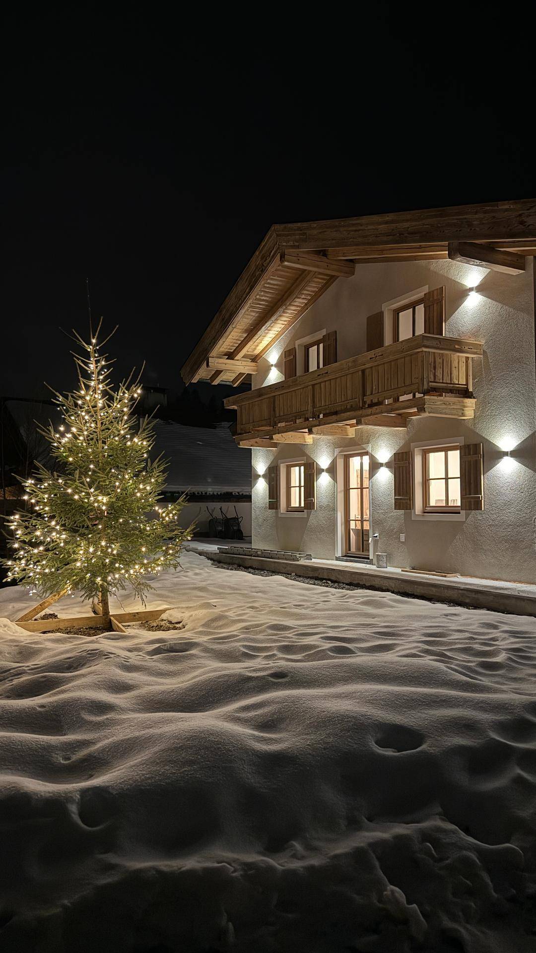 Alpines Haus bei Nacht im Schnee, festlich beleuchtet mit einem geschmückten Weihnachtsbaum.