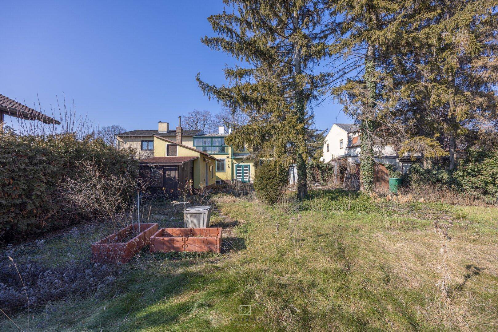 Gartenbereich mit Hochbeeten und Blick auf die Rückseite des Hauses unter blauem Himmel.