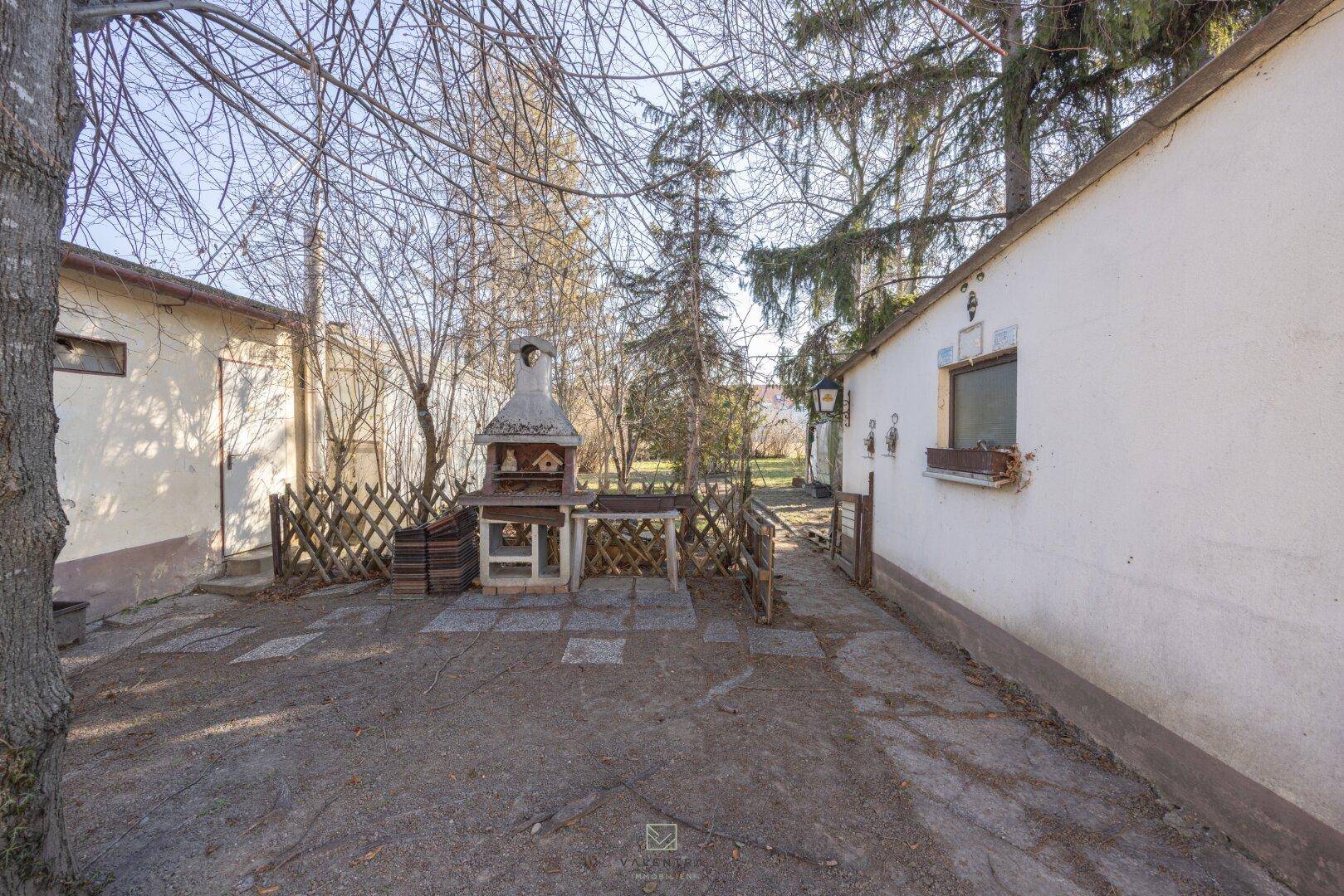 Außenbereich mit einem gemauerten Grillplatz und einer gepflasterten Fläche zwischen zwei Gebäuden.
