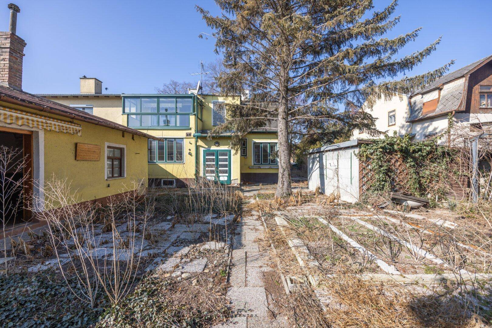 Rückseite eines gelben Hauses mit Wintergarten und verwildertem Gartenbereich.