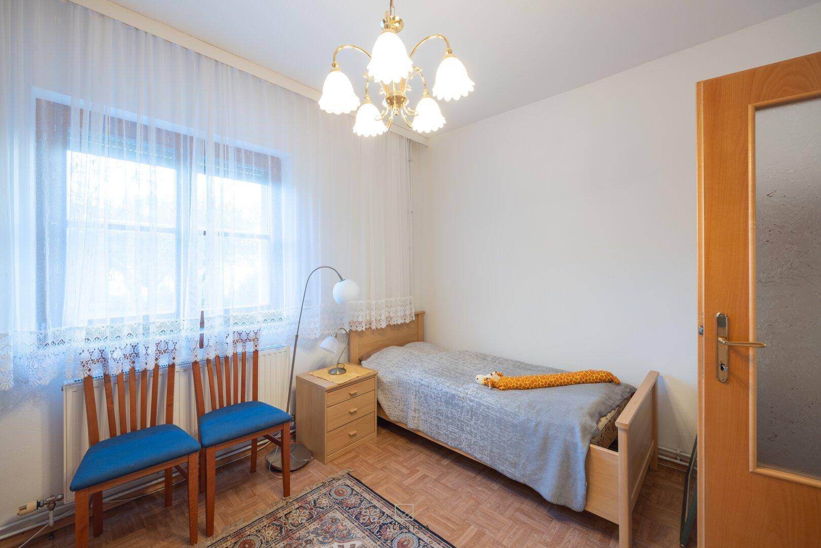 Helles Schlafzimmer mit Einzelbett, Holzmöbeln und Fenster mit Vorhängen.