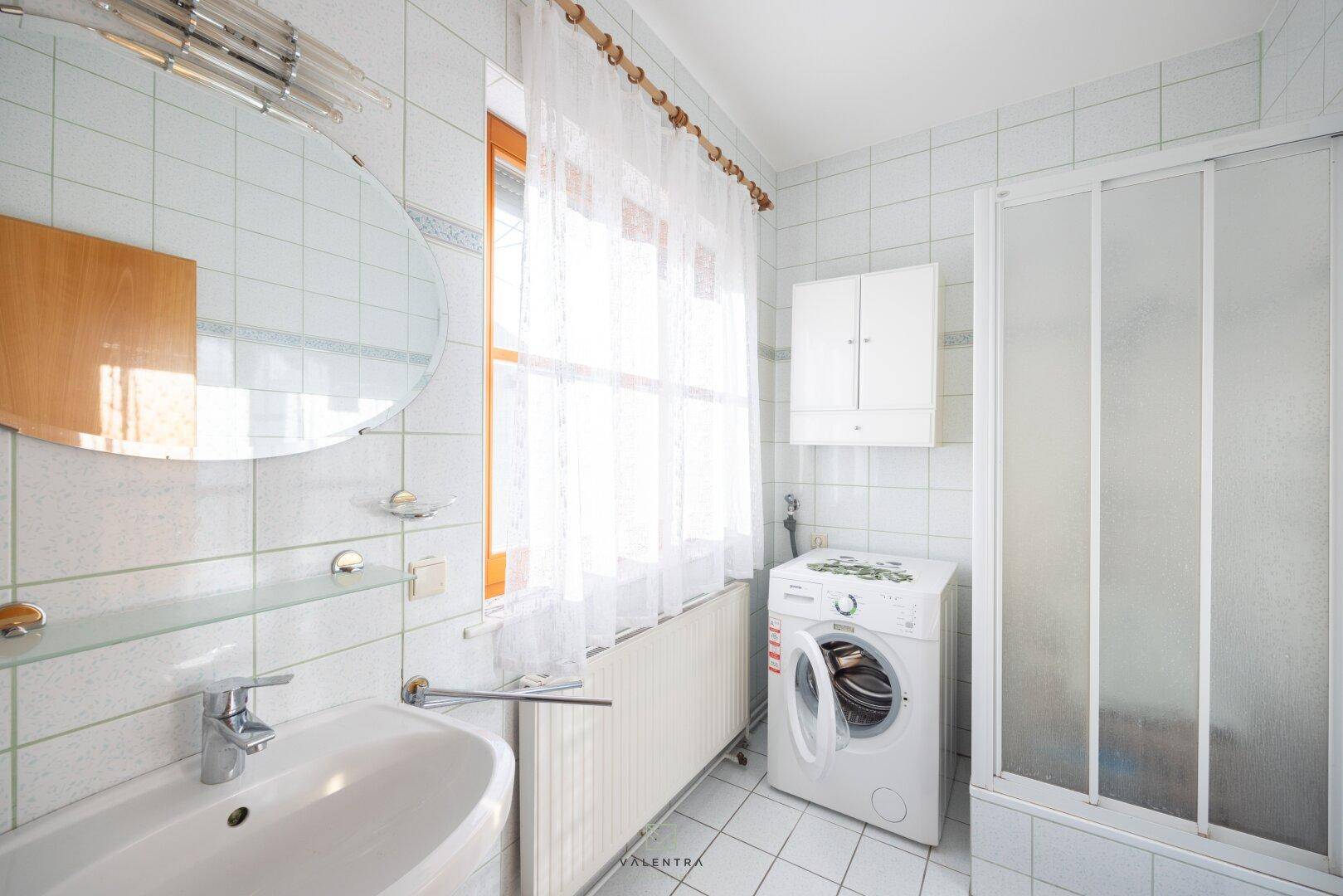 Helles Badezimmer mit Dusche, Waschbecken, Waschmaschine und Fenster.