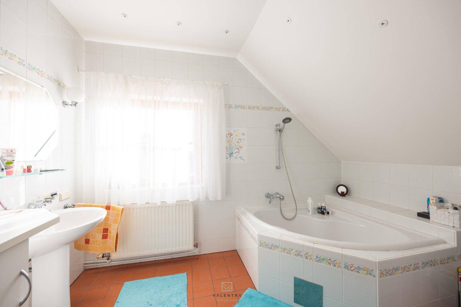 Badezimmer mit Eckbadewanne, Waschbecken und Fenster unter der Dachschräge, gefliestem Boden.
