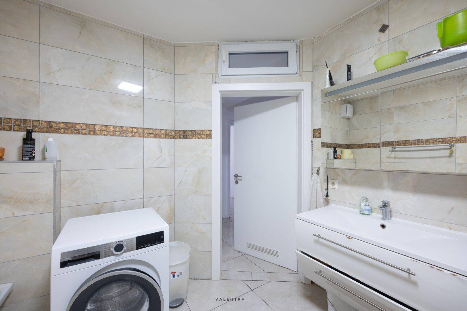 Badezimmer mit Waschmaschine, Waschbecken und hellen Fliesen, inklusive Fenster für Tageslicht.