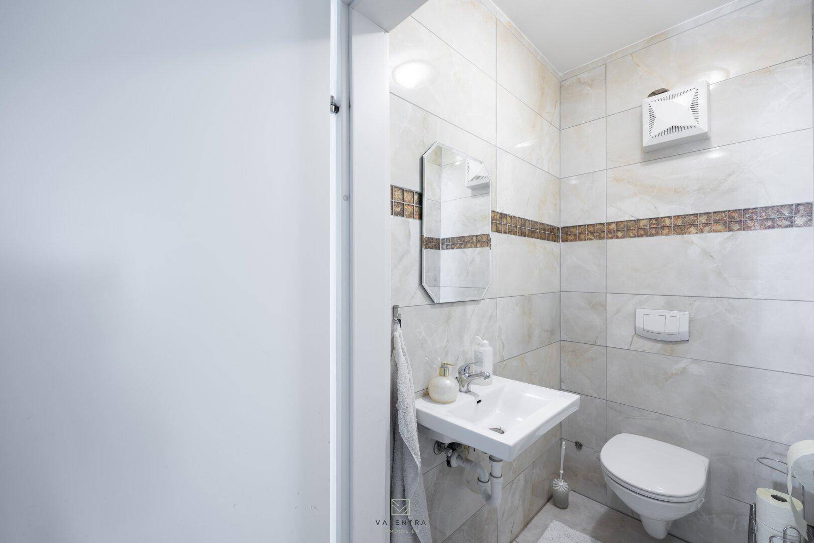 Kleines Gäste-WC mit Toilette, Waschbecken und Spiegel, ausgestattet mit hellen Fliesen.
