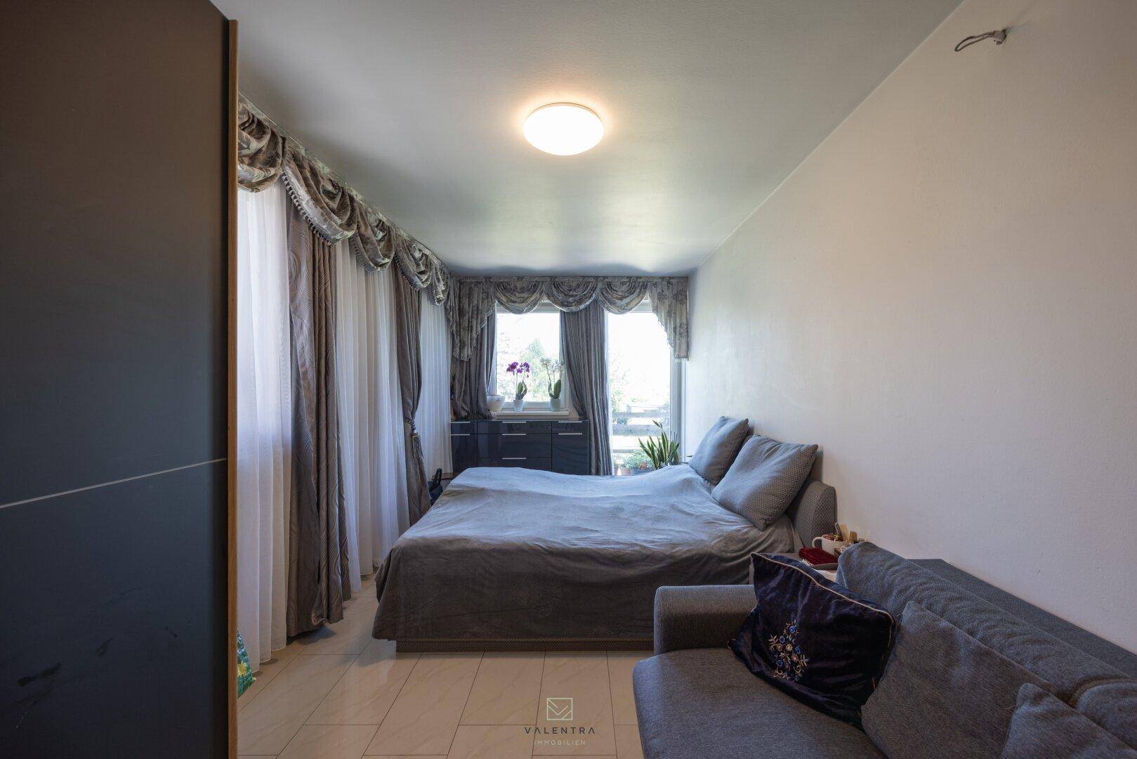 Gemütliches Schlafzimmer mit Bett, Sofa und Fenster, geschmückt mit eleganten Vorhängen.