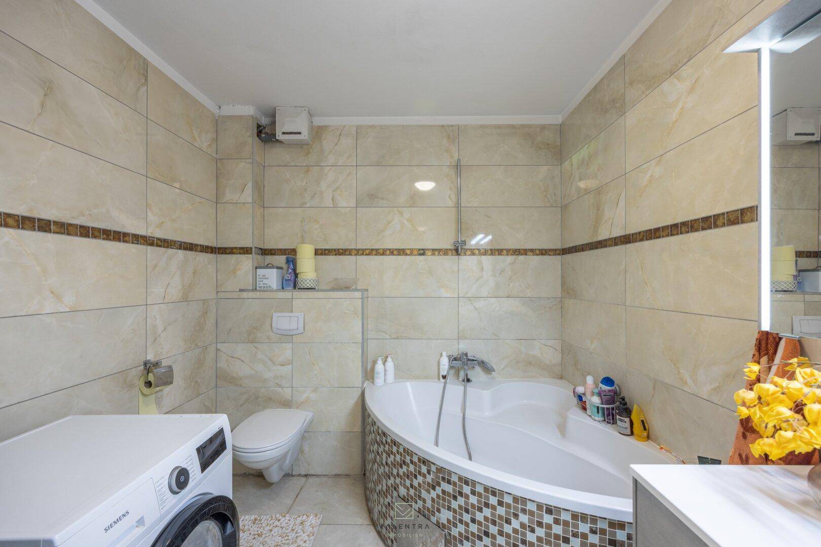 Badezimmer mit Eckbadewanne, Toilette und Waschmaschine, komplett gefliest in hellen Farbtönen.