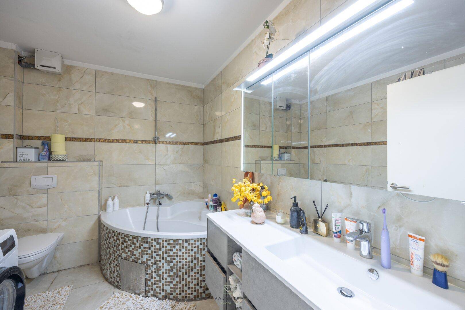 Helles Badezimmer mit Eckbadewanne, Doppelwaschbecken und großem Spiegelschrank, vollständig gefliest.