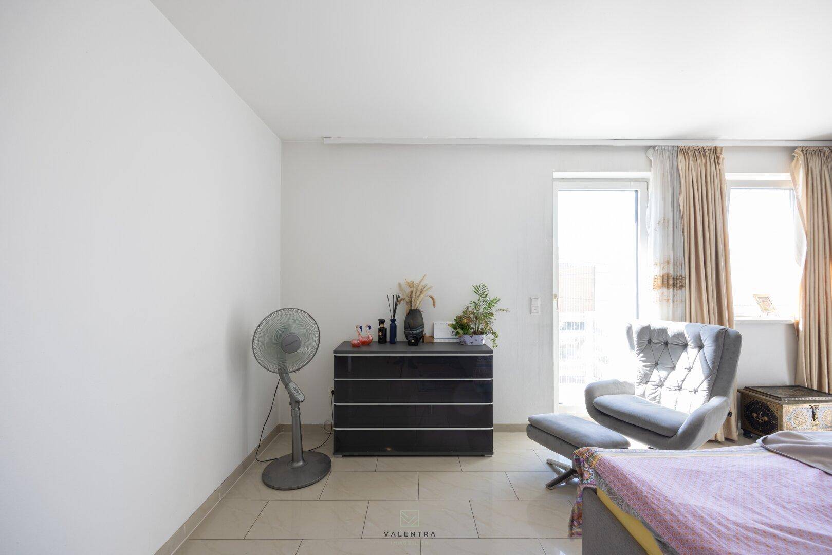 Schlafzimmer mit Bett, Kommode und Sessel, hell durch Fenster mit Vorhängen, auf Fliesenboden.